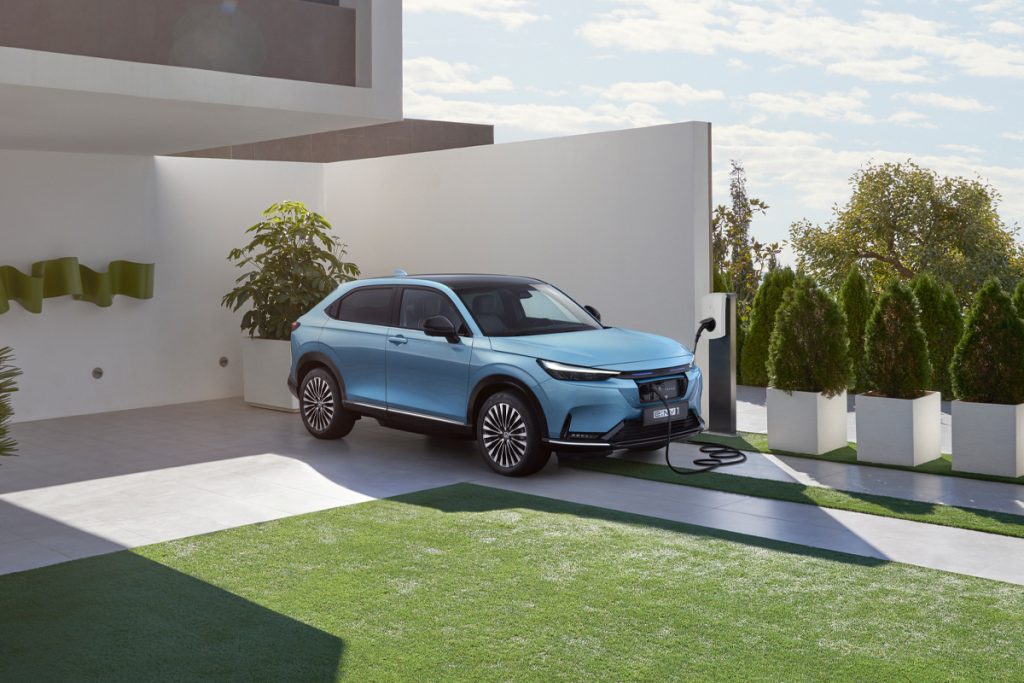 A front right view of a Honda e:Ny1 plugged in to an Alfen S-Line charger in a picturesque villa setting